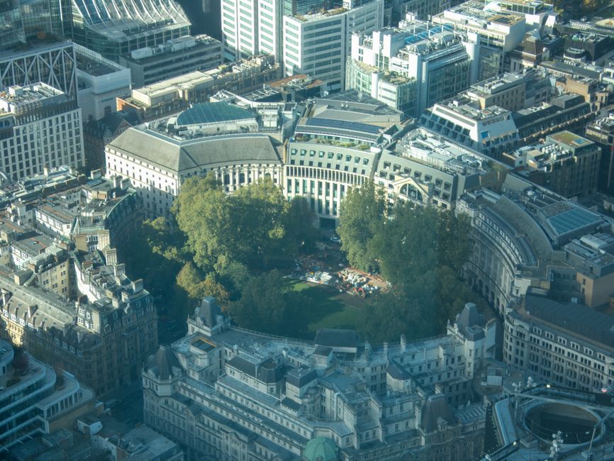 Aerial view of a cityscape featuring a circular park with lush green trees at its center, surrounded by historic and modern buildings with varied architectural styles. The park area includes visible p