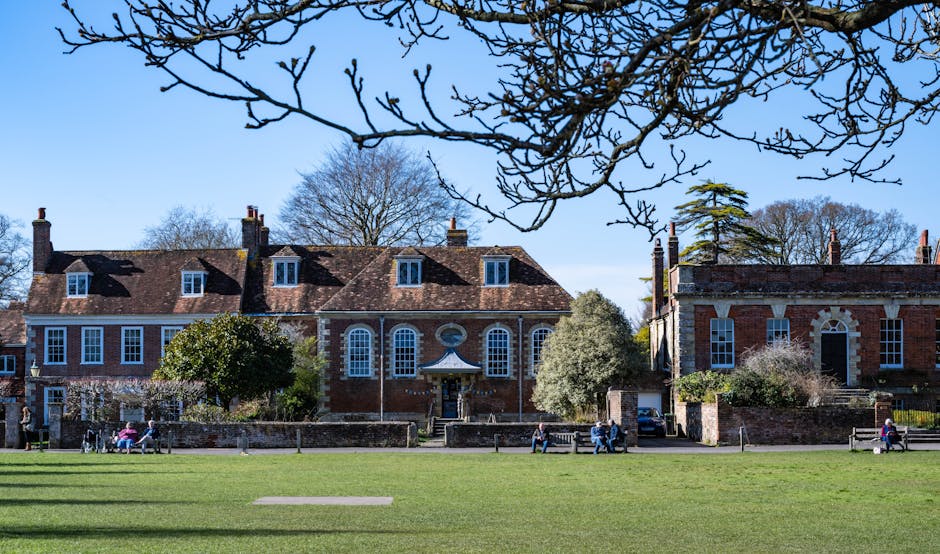 View of a historic red brick building with white-framed windows and a central entrance, set against a background of leafless trees under a clear blue sky. The foreground features a well-maintained gre