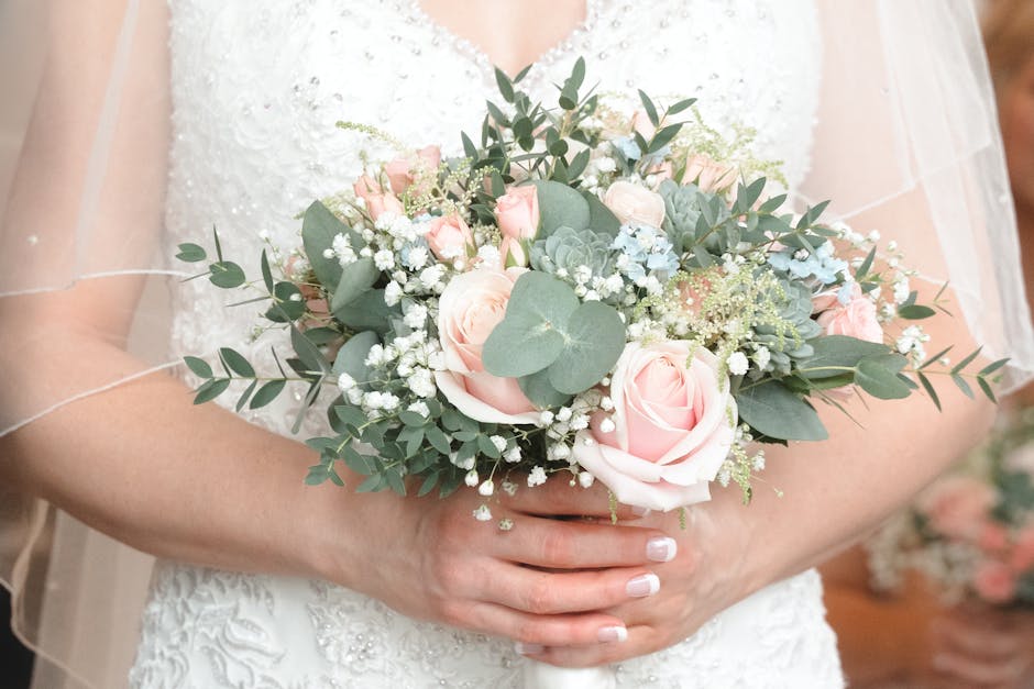 A bridal bouquet featuring soft pink roses, white baby's breath, and pale blue hydrangeas, accented with eucalyptus leaves and succulents, held by a bride in a lace wedding dress. The arrangement is l