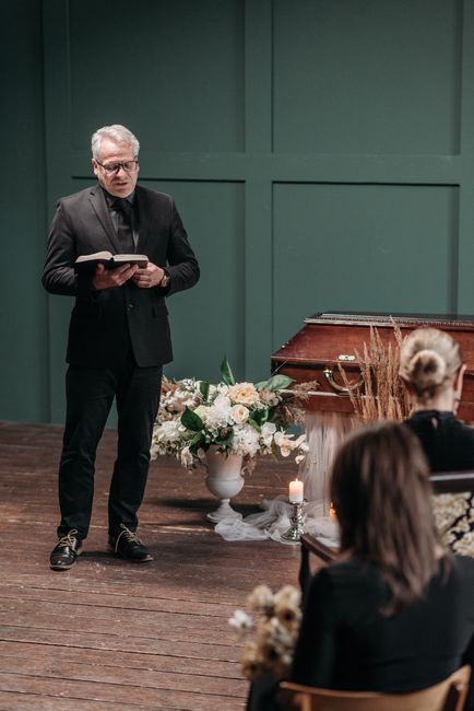 A man dressed in a black suit with glasses stands on a wooden stage, holding an open book, delivering a speech at a funeral ceremony. In front of him is a large floral arrangement in a white vase, fea