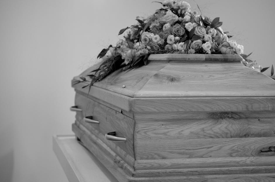A wooden casket with a smooth, polished surface and natural wood grain visible, topped with a floral arrangement of mixed flowers including roses, lilies, and carnations in shades of white, cream, and